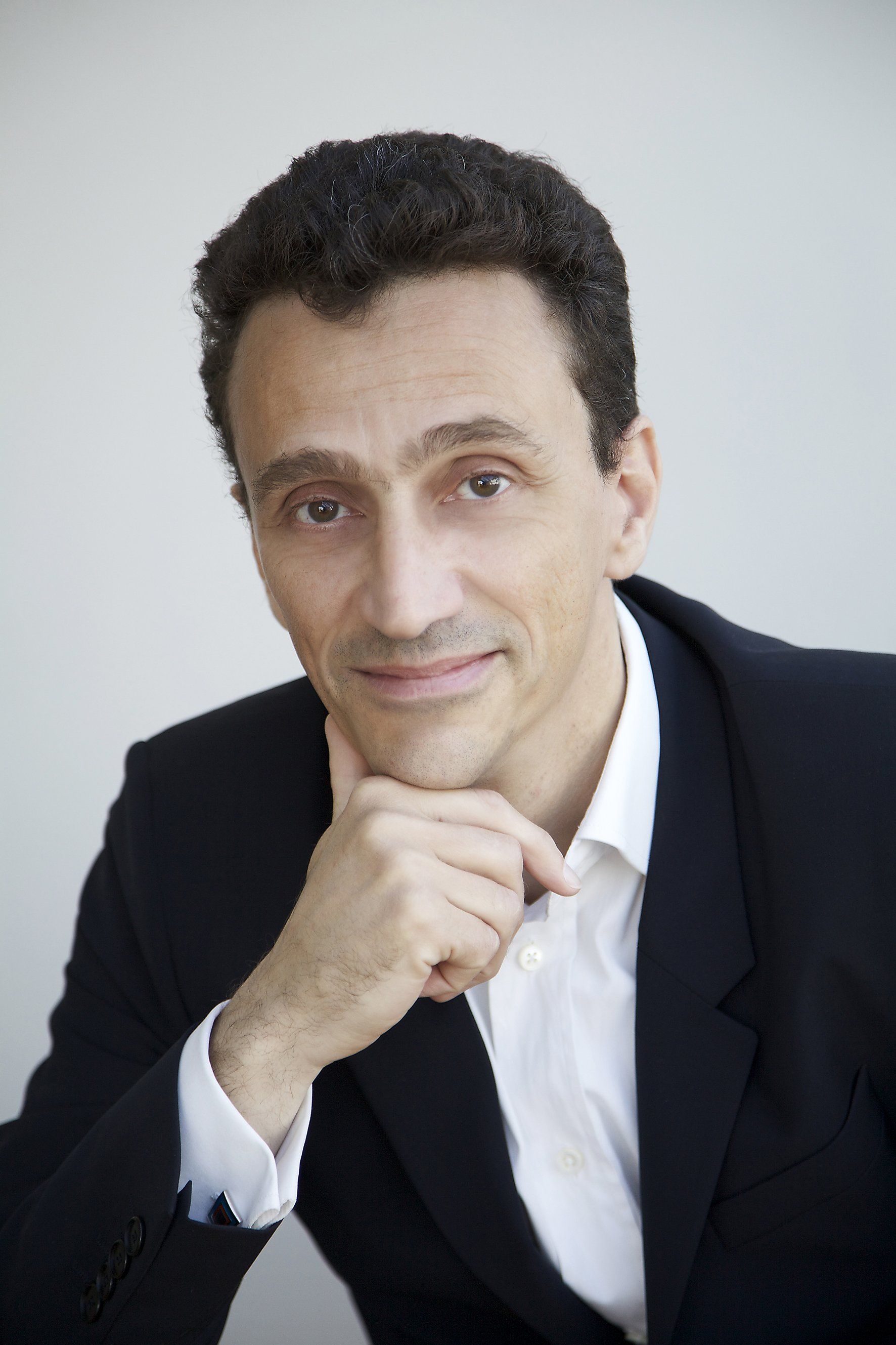 Portrait d'un homme en blazer foncé et chemise blanche, le menton appuyé sur sa main, sur fond clair.
